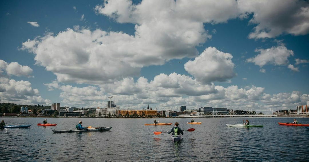 Stadssiluett och bl&aring; himmel med vita moln och en sj&ouml; i f&ouml;rgrunden d&auml;r n&aring;gra paddlar kajak.